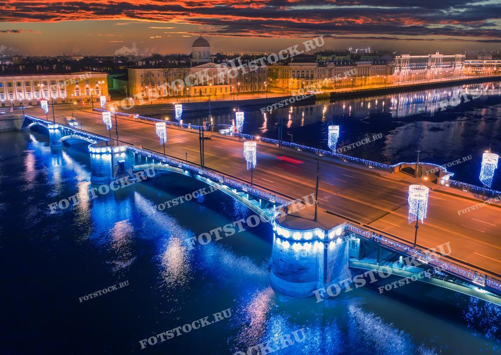 На фотографии изображён вечерний Санкт-Петербург, украшенный праздничной иллюминацией. Световые гирлянды на мосту и его отражение в воде создают праздничное и торжественное настроение. 
Это изображение идеально подходит для использования в туристических путеводителях, рекламных материалах, посвящённых Санкт-Петербургу, а также в образовательных и культурных публикациях.